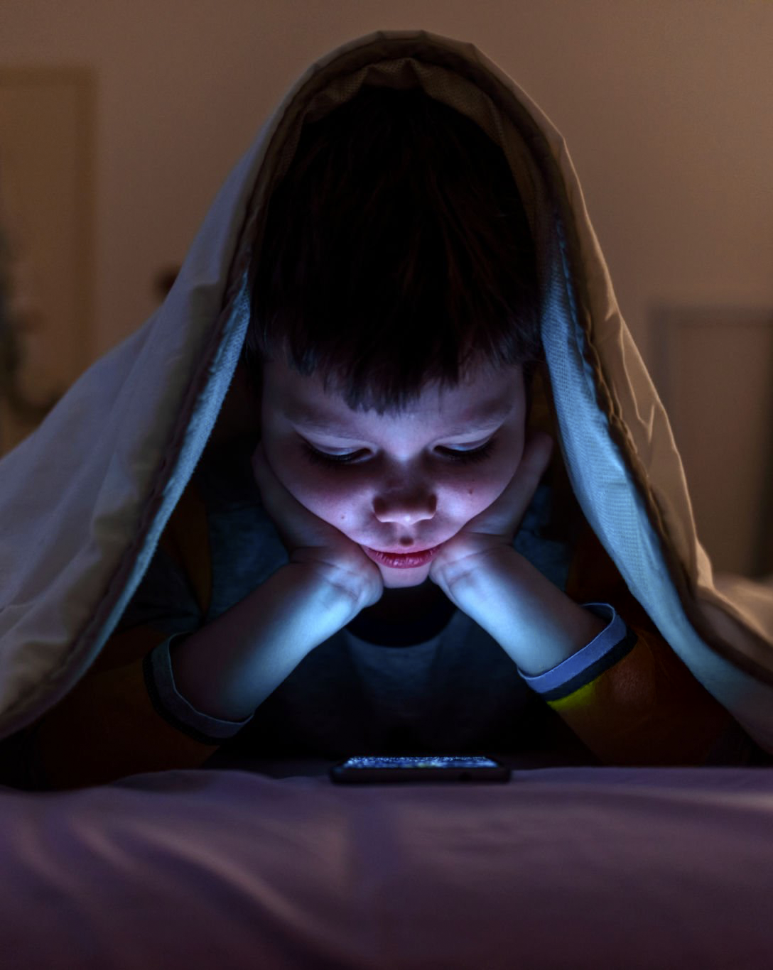 Criança sozinha no escuro olhando o celular embaixo do lençol